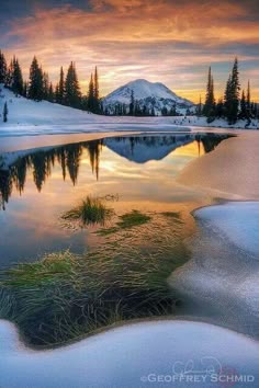 a lake surrounded by snow covered mountains and evergreen trees at sunset with the sun setting in the distance