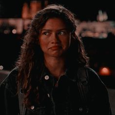 a woman with long hair standing in front of a building at night, looking off into the distance