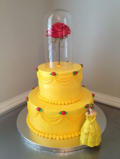 a yellow cake with a red rose in a glass dome on top and a small figurine next to it