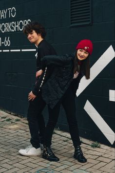 two people standing next to each other on a brick sidewalk in front of a black wall