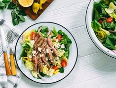 two plates of salad with meat and vegetables next to each other on a white table