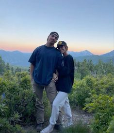 a man and woman standing on top of a lush green hillside with mountains in the background