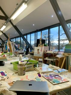 an artist's studio with lots of paintings and art supplies on the table in front of large windows