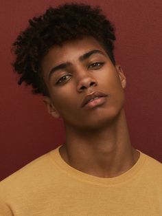 a young man with curly hair wearing a yellow t - shirt and looking at the camera