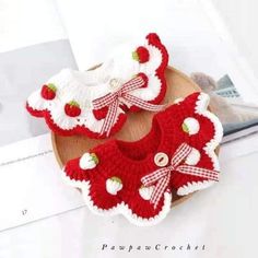 two red and white crocheted baby booties with bows on the side, sitting on top of a wooden plate