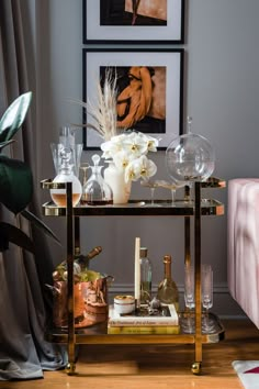 a bar cart with wine glasses, bottles and flowers on it in front of two framed pictures