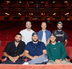 five men are sitting in the middle of an empty auditorium with red seats and one man is smiling at the camera