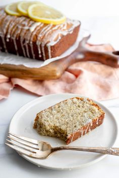 a slice of lemon cake on a white plate with a fork next to it and a loaf of lemon cake in the background