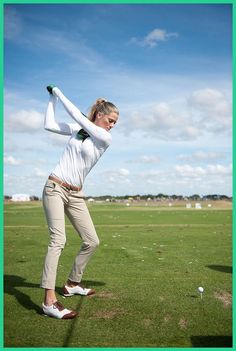 a woman in white shirt and tan pants swinging a golf club on green grass field