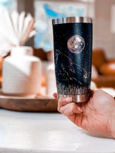 a person holding up a travel mug with the moon and stars in the sky on it