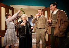 four people standing in a living room with one woman holding a wine glass and the other man reaching for something