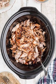 shredded meat in a slow cooker on a white wooden table with utensils