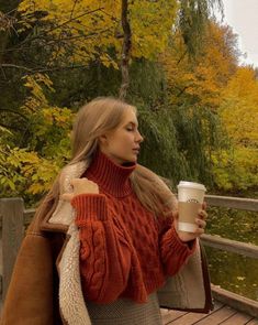 a woman holding a coffee cup while standing on a wooden bridge in the fall time