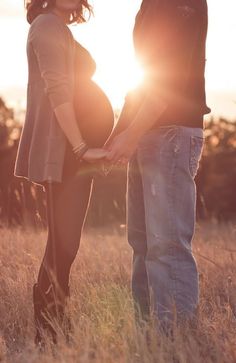 a pregnant couple holding hands while standing in tall grass at sunset with the sun behind them