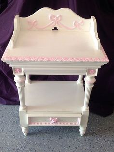 a white and pink baby changing table with bow on it's top, in front of a purple background