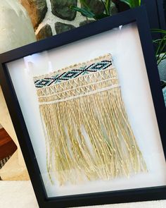 an art piece with fringes is displayed in a black frame on a table next to a potted plant