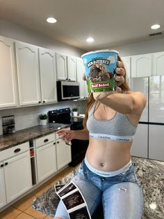 a woman sitting on top of a kitchen counter holding up an ice cream container in front of her face