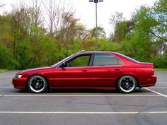 a red car parked in a parking lot next to some trees and bushes on the other side