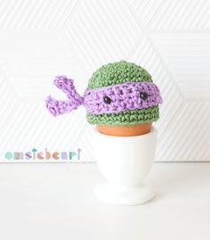 there is a crocheted hat on top of an egg shell with a purple and green decoration