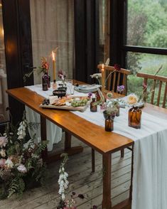 a table with flowers and candles on it in front of a large window, next to a wooden bench