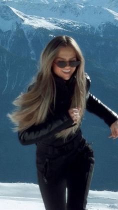 a woman with long blonde hair and sunglasses on top of a snow covered slope in the mountains