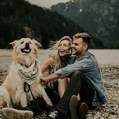 a man and woman sitting next to a dog on the ground with mountains in the background