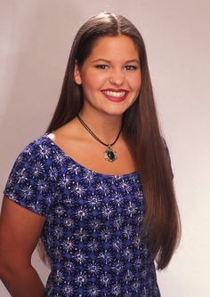 a woman with long brown hair wearing a blue dress and smiling at the camera while standing in front of a white wall