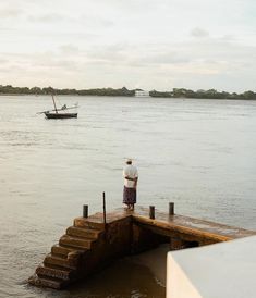 a man standing on a dock watching a boat in the water and another person with a hat