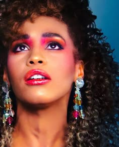 a woman with long curly hair and bright makeup looks into the distance while wearing colorful earrings