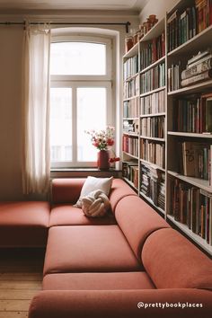 a red couch sitting in front of a book shelf filled with lots of books next to a window