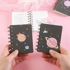 two hands holding notebooks with planets and stars on them in front of a pink background