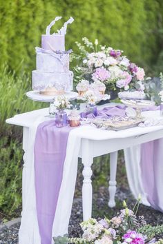 a table topped with a purple cake and lots of flowers next to a lush green forest