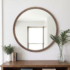 a round mirror on the wall above a wooden shelf with books and plants in it