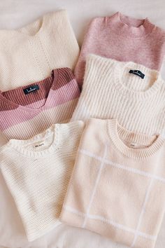 three sweaters laying on top of a bed with white sheets and pink, beige, and blue colors