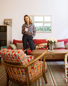 a woman standing in a living room next to a red couch and chair with pillows on it