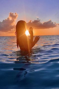 a woman sitting in the water with her hands up to her face as the sun sets