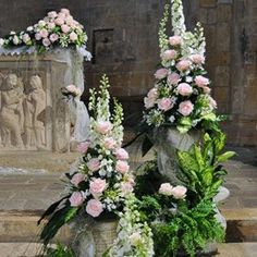 two vases with flowers and greenery on the side of a stone pillar in front of a statue