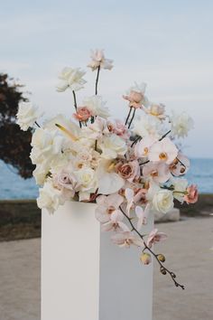 a white vase filled with lots of flowers on top of a sandy beach next to the ocean