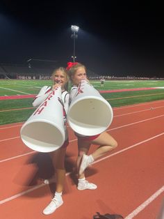 two girls in cheerleader outfits holding large white buckets on a track at night