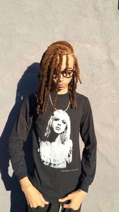 a young man with dreadlocks standing against a wall wearing a t - shirt