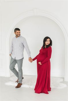 a pregnant woman in a red dress holding the hand of her husband as they walk through an archway