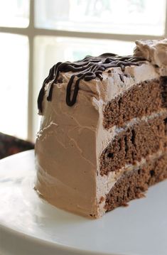 a slice of chocolate cake with frosting on a white plate in front of a window