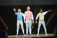 three young boys in colorful vests and white pants are on stage with their hands up