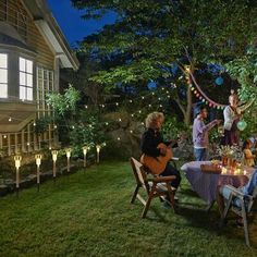 a group of people sitting around a table outside at night with lights on the trees