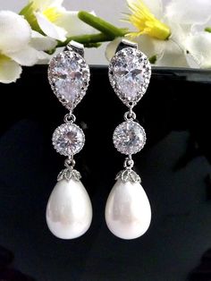 two pairs of white pearl and crystal earrings on a black surface with flowers in the background