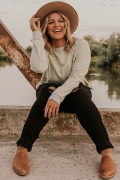 a woman sitting on the edge of a bridge wearing a brown hat and black pants