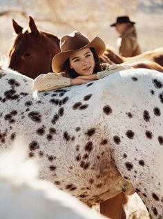 a woman wearing a cowboy hat standing next to a spotted horse in the middle of a field