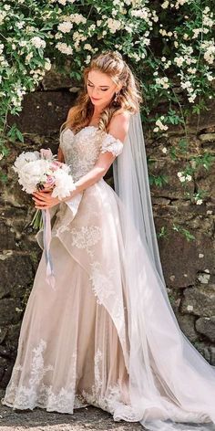 a woman in a wedding dress holding a bouquet of flowers and ribbons next to a stone wall