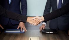 two people shaking hands over a table with other people in suits and ties behind them