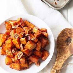 a white plate topped with sweet potatoes next to a wooden spoon and bowl of nuts